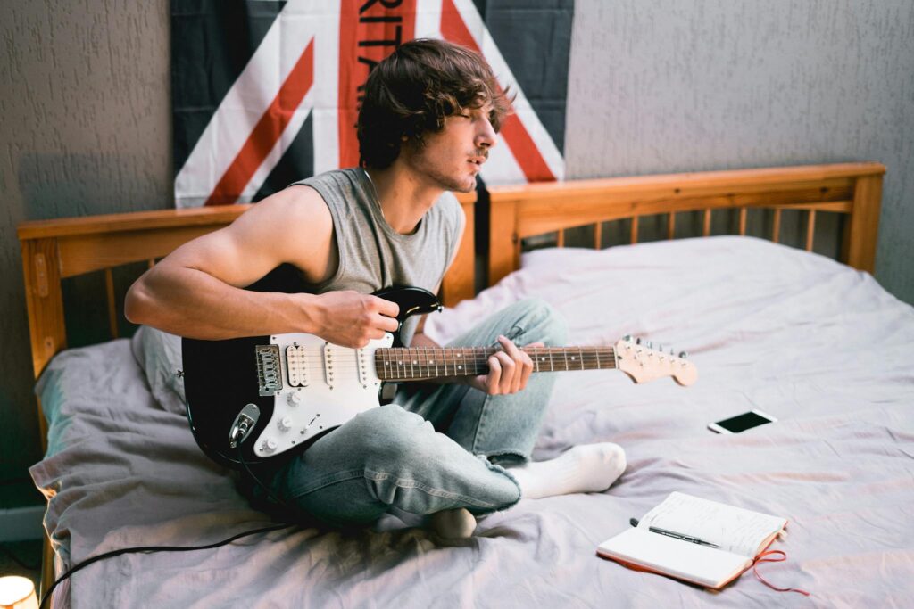 A young musician sits on a bed with a guitar, composing music and writing in a notebook.