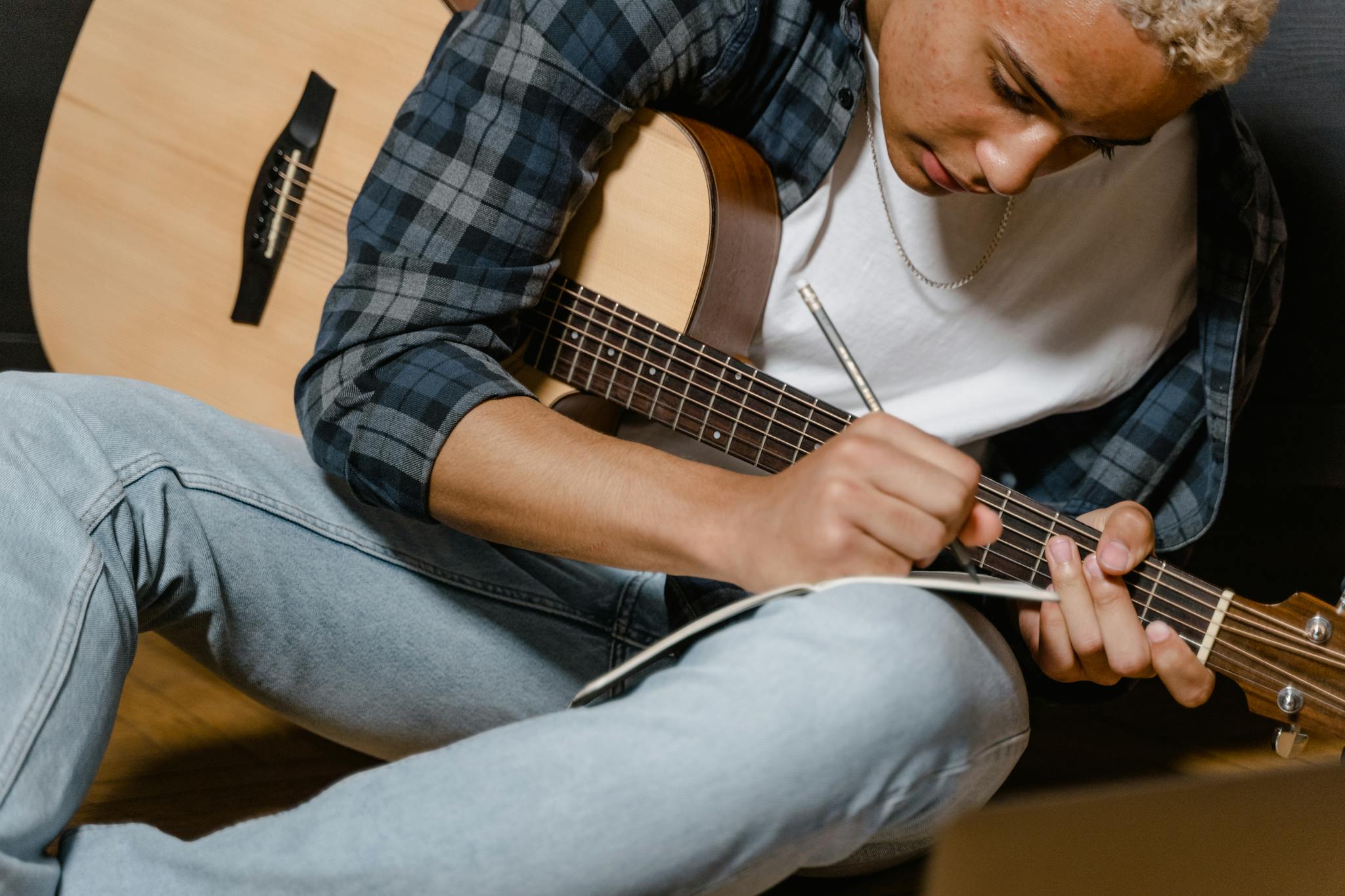 A young musician writes lyrics with a guitar by his side, deep in thought.