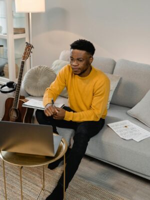Man in yellow sweater composing music with guitar and laptop in a modern living room.