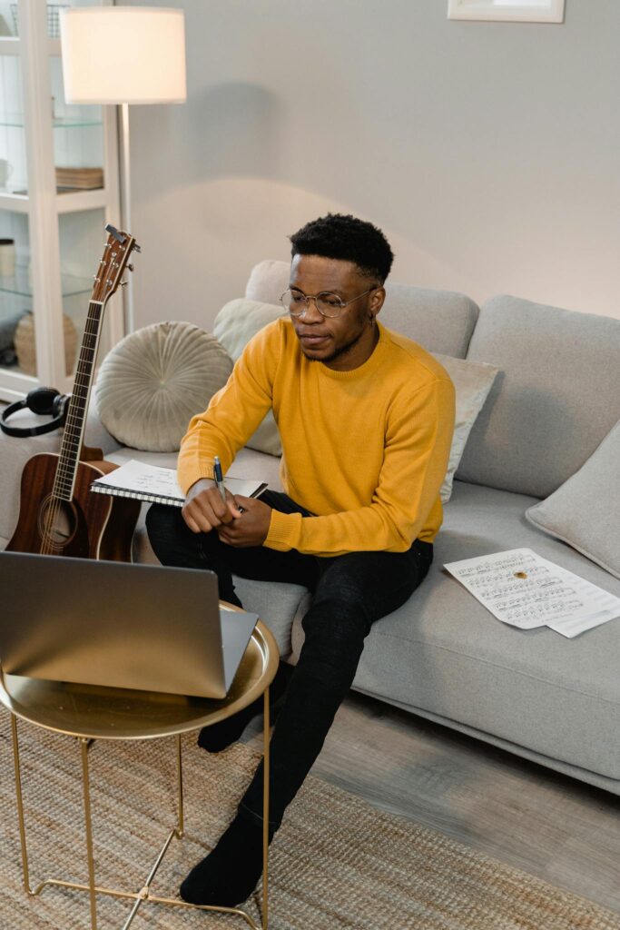 Man in yellow sweater composing music with guitar and laptop in a modern living room.