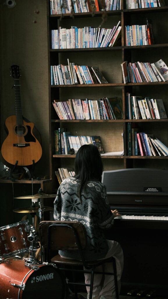 Woman playing piano in a library with guitar and drums nearby, evoking a creative atmosphere.